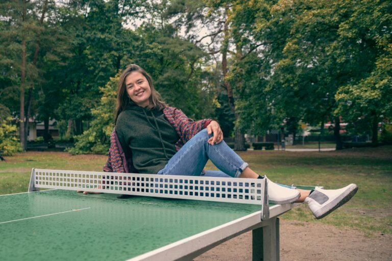 A cheerful woman poses on a ping pong table outdoors in a park setting, surrounded by lush greenery.